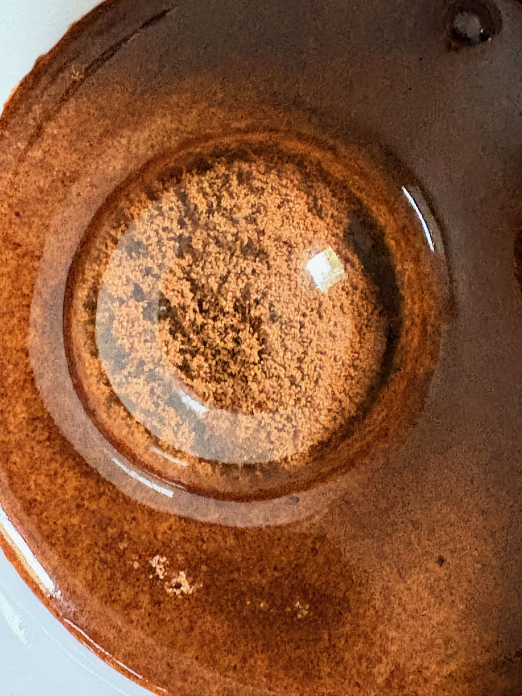 Vista dall'alto di una superficie ceramica smaltata color ruggine: una cupola d'acqua trasparente copre uno strato granuloso di caffè solubile.